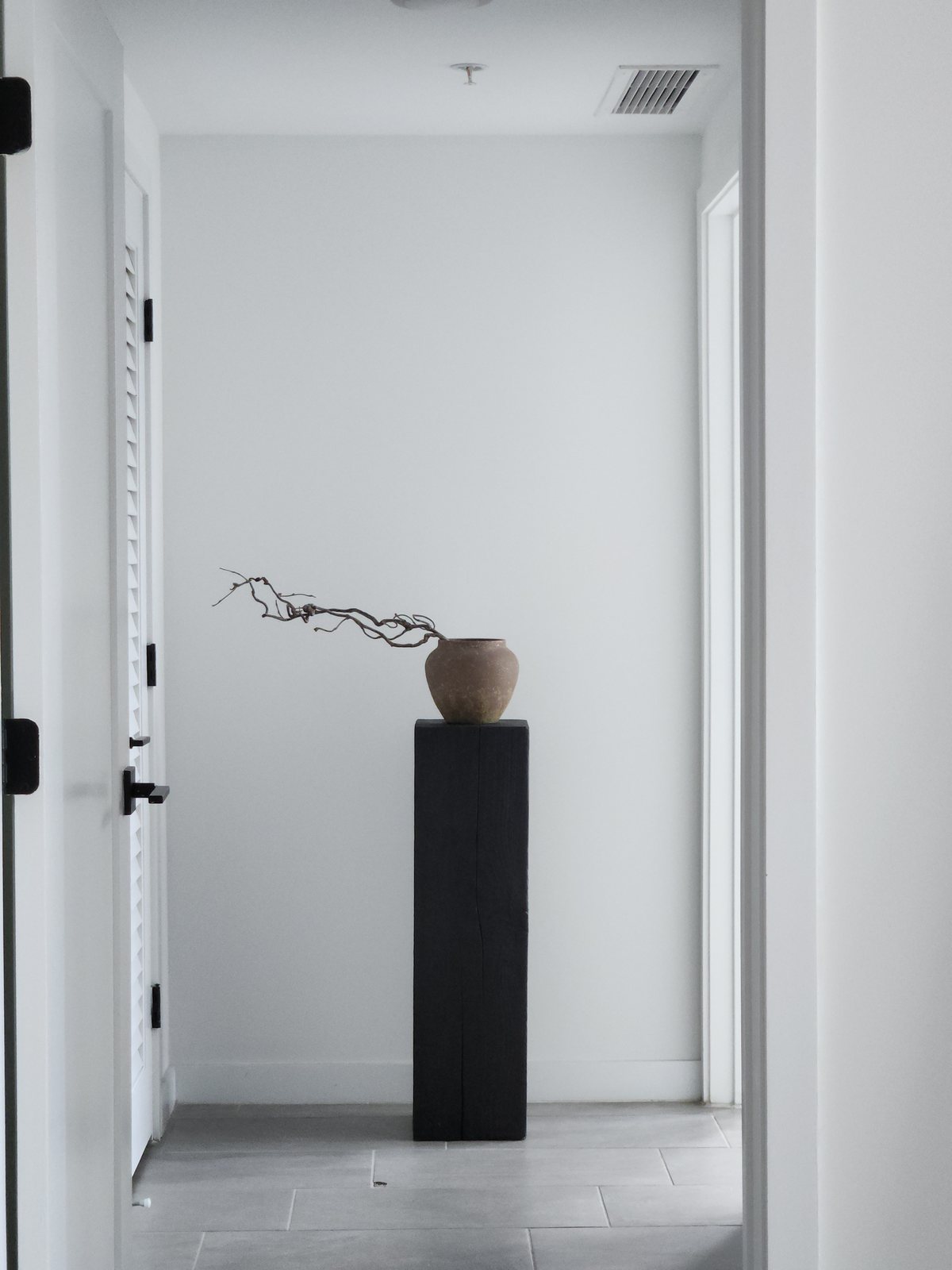 Black pedestal with a clay vase and branch at the end of a white hallway