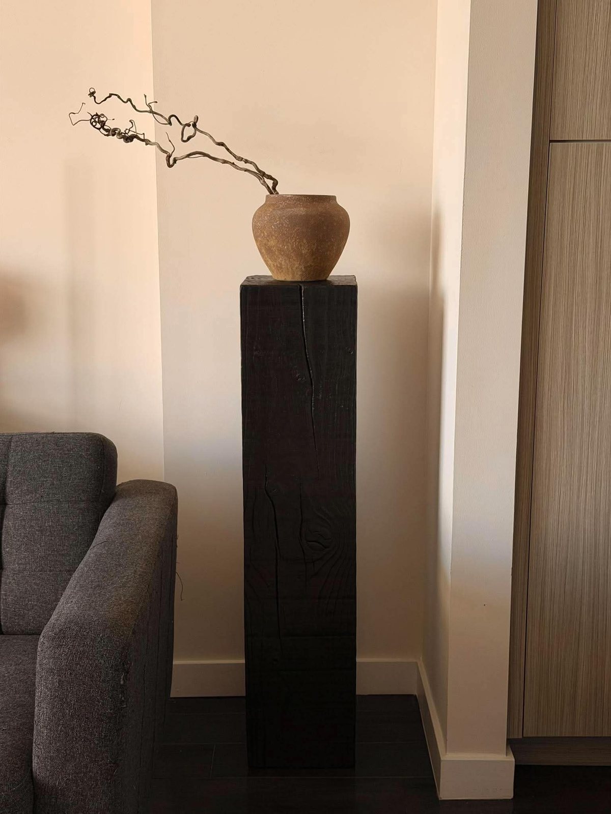 Tall black pedestal with a clay vase next to a grey sofa