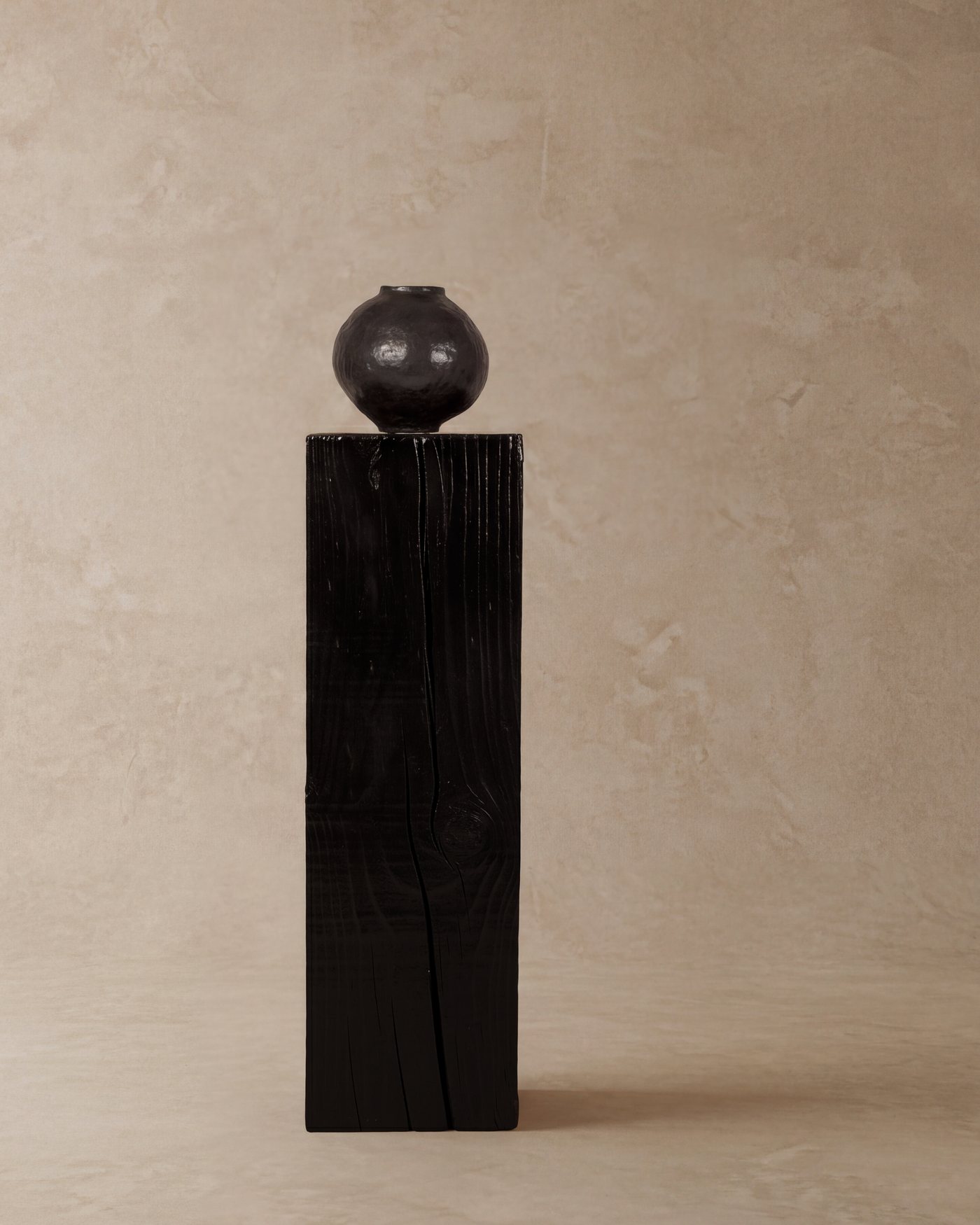 Black charred pedestal with a dark round vase against a plaster backdrop